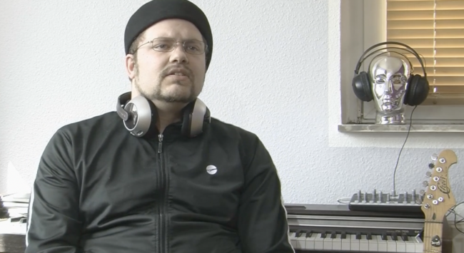 Person mit Kopfhörern um den Hals, E-Piano im Hintergrund und Gitarrenkopf daran gelehnt