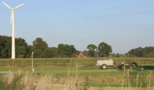 Landschaft mit Windrad und Feld und Trekker mit Güllewagen dahinter Ziegelhaus
