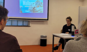Seminarraum mit Vortragender und Präsentationsslide auf dem steht Mallorca Deutsche Ausgabe und Rücken von Zuhörenden im Vordergrund